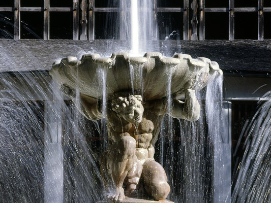 Eine klassizistische Statue stemmt eine muschelförmige Schale, in die Wasser prasselt. Wasser läuft zu den Seiten heraus. Eine Nahaufnahme eines antiken Brunnens.